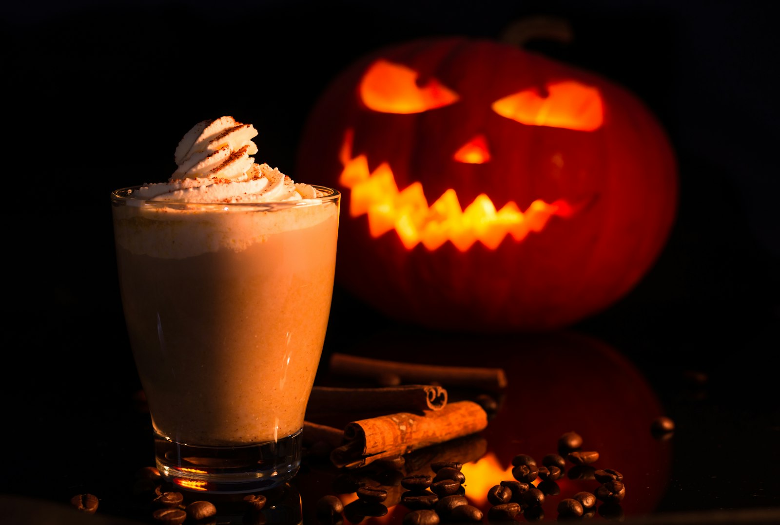 A pumpkin spice latte topped with whipped cream beside a glowing jack-o-lantern on a dark wooden table